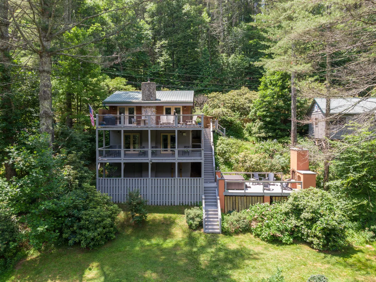 A spacious, multi-level deck surrounded by lush greenery, a stone chimney, and a flagpole, set up for gatherings and outdoor relaxation. The scene is tranquil and welcoming, with potential for evening fireside moments.