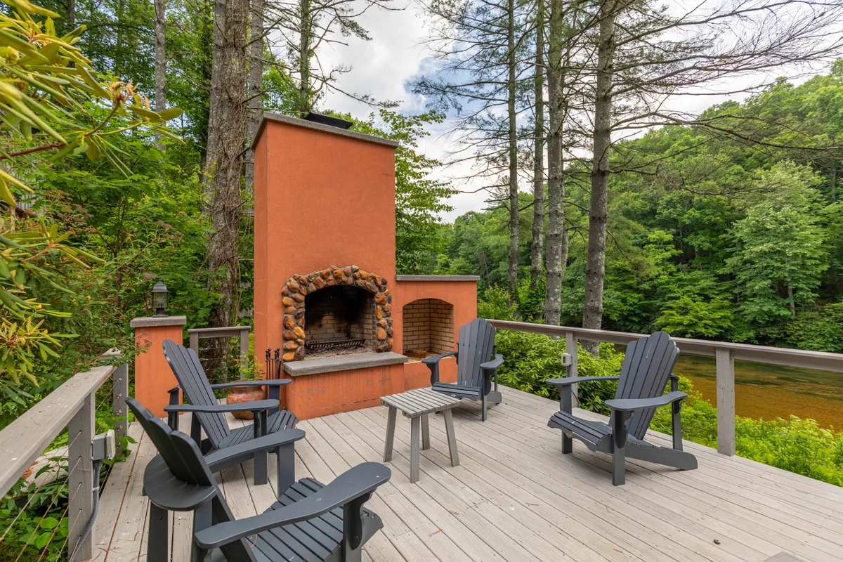 This image showcases the private wooden deck, featuring a substantial stone outdoor fireplace, comfortable chairs, and lush greenery all around. The setting appears tranquil and inviting, perfect for relaxing by the fire or gathering with friends and family against a beautiful natural backdrop.