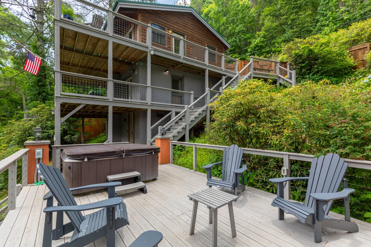 A spacious deck area features an inviting hot tub and Adirondack chairs, all set amid greenery beside the house. The river is visible beyond, offering a seamless connection between relaxation, socializing, and the outdoors.
