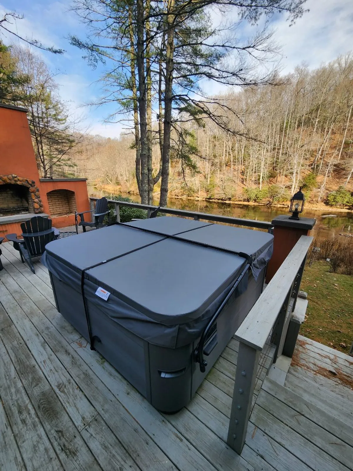 An inviting exterior view showing a private deck with a hot tub, comfortable seating, and a lush river view beyond. The space is surrounded by trees, offering seclusion and direct access to natural beauty.