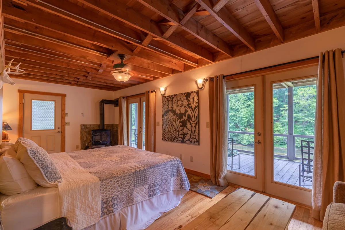 A tranquil master bedroom with a bed adorned with a decorative quilt, large windows framing views of nature, a fireplace for added coziness, and artwork on the wall. The wooden ceiling enhances the warmth and retreat-like ambiance.