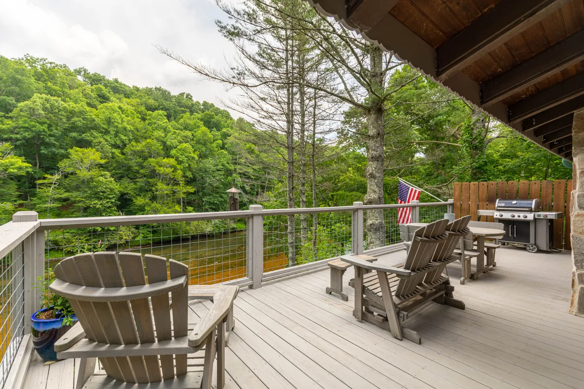 A spacious wooden deck with lounge chairs, grill, and river views, surrounded by mature trees. The space overlooks the water and provides a comfortable spot for relaxing, socializing, and soaking in the outdoors after a day of adventures.