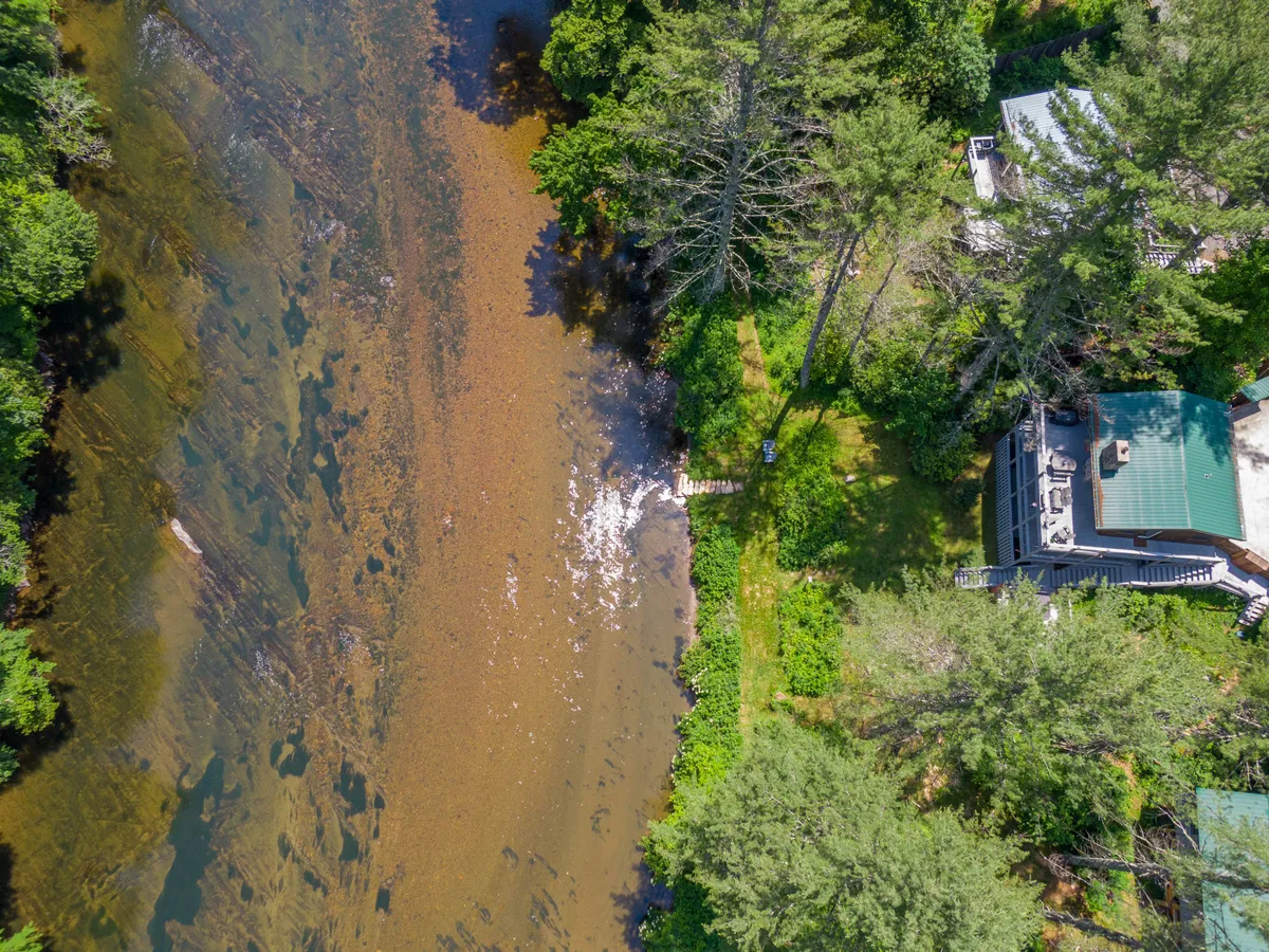 A wide exterior shot showing the riverside setting with the house, a dock extending into the water, mature trees, and an inviting atmosphere. The scene captures both the waterside access and the natural surroundings.