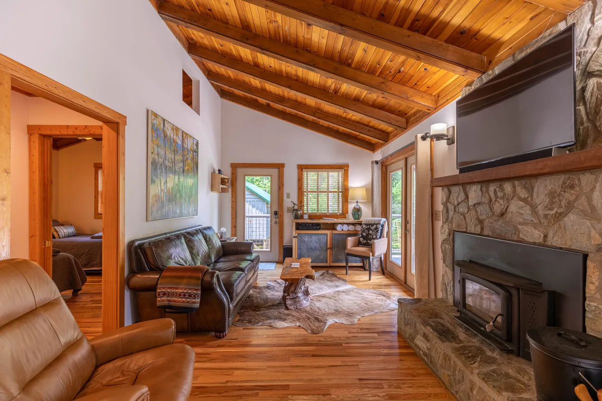 A cozy living room featuring a stone fireplace, leather sofa, wooden beams overhead, and a rustic coffee table. Natural light pours in through generous windows, illuminating the comfortable seating area and mountain-inspired artwork on the walls.