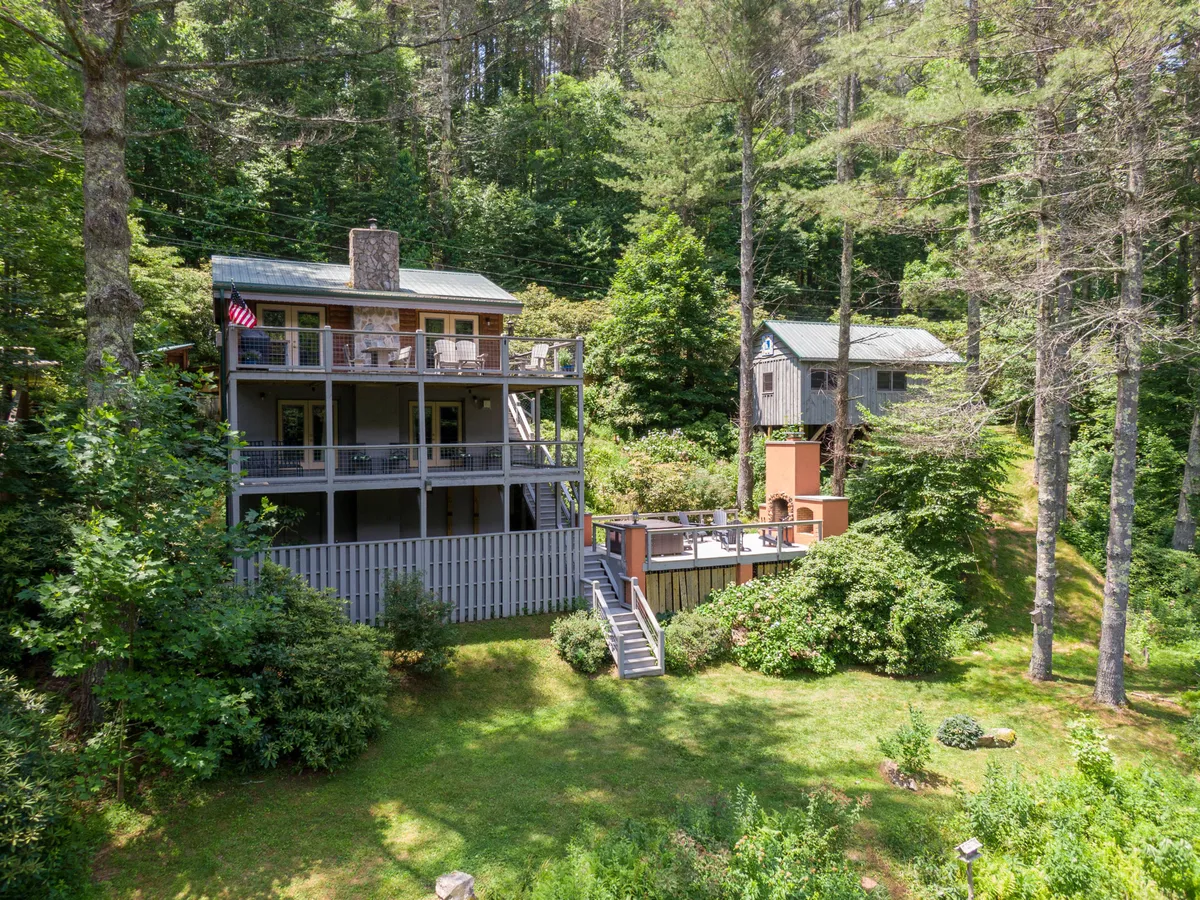 This image features a charming two-story mountain cabin nestled among mature trees, with a spacious deck furnished for outdoor relaxation. The inviting garden area and serene natural surroundings highlight the property's tranquil, riverfront setting, perfect for sunset views and unwinding close to nature.