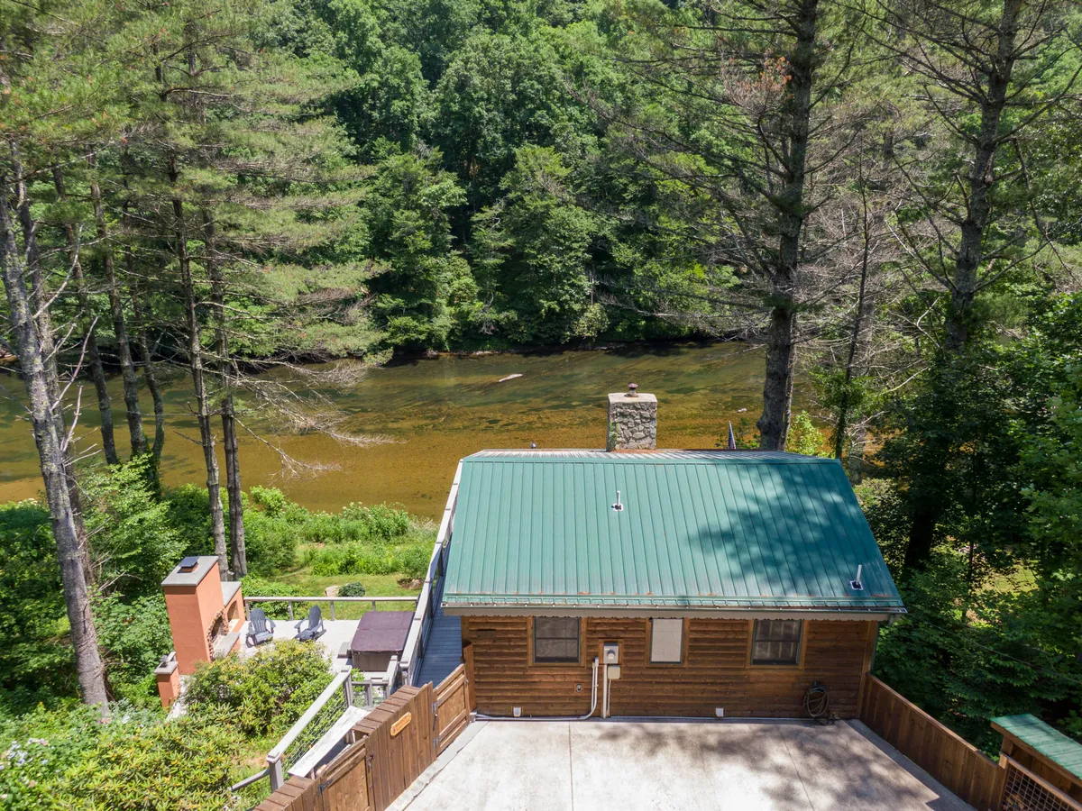 A charming log cabin with a green roof sits nestled among tall trees, right beside the gentle flow of the New River. The setting is serene, with a sense of seclusion, and the riverbank offers direct access to nature trails and water activities. The image captures both the inviting home and the natural amenities ideal for outdoor adventures with pets.